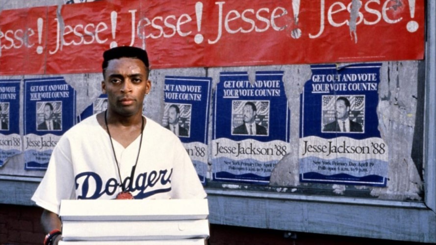 A man in a Dodgers jersey holds three pizza boxes. He is looking directly to camera. Behind him are posters for Jesse Jackson '88.