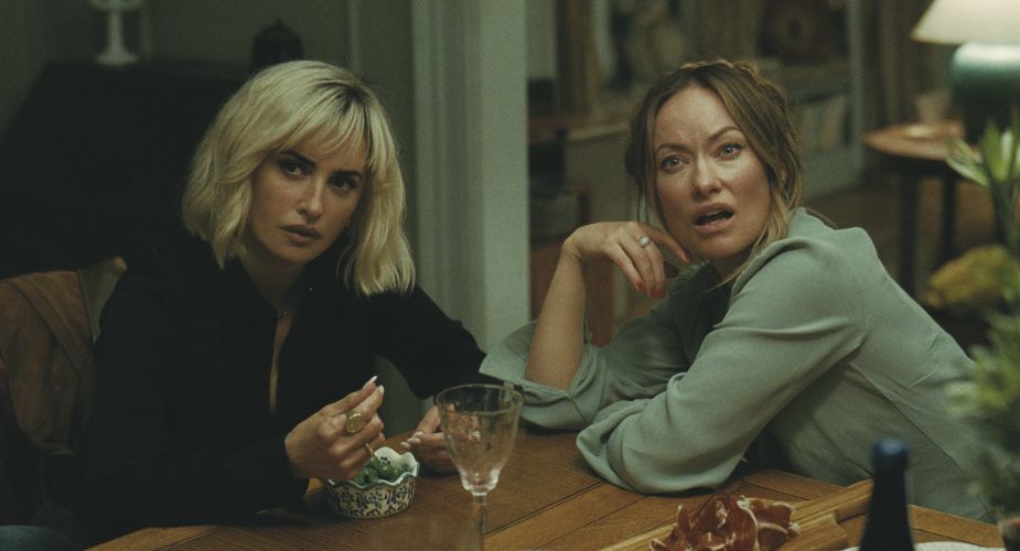 Two women sit at a table, one woman with a short blonde bob looks off-camera with a look of contempt. The other woman in a green shirt looks in a similar direction, disgusted.