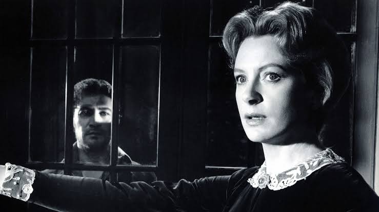 A black and white image from The Innocents. A woman in the foreground, looking concerned away into the distance, a man stands behind her on the other side of a window.
