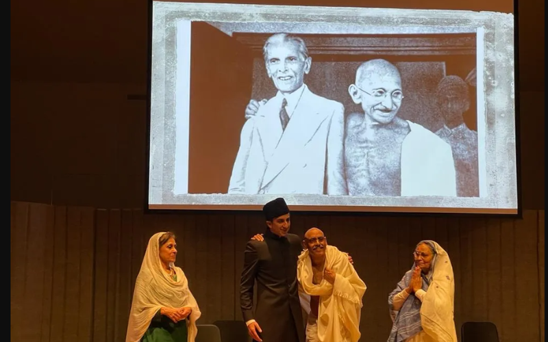 Two women actors, dressed in Saris, look on at two men who are dressed as Gandhi and Jinnah. Behind them, a photograph of the real Gandhi and Jinnah.