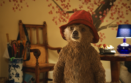 A still from Paddington showing the bear wearing his little red hat standing in the entrance to the Brown's house.