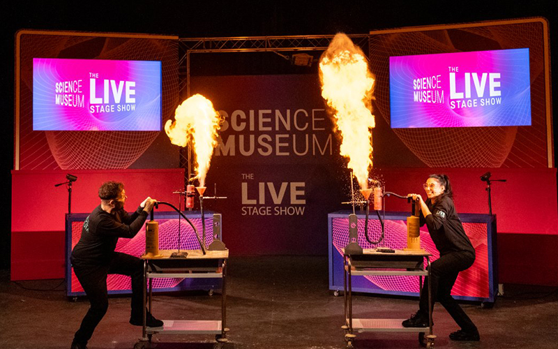 Two people on stage as part of Science Museum The Live Stage Show. They are both standing on trolleys and causing fire to come out of large Bunsen burners (they are wearing safety goggles).