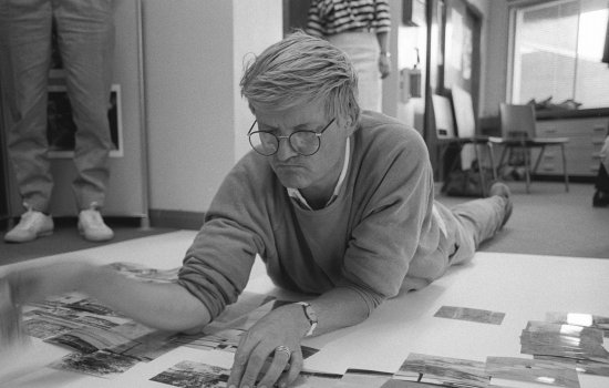 A photograph of David Hockney lying down arranging photos on the floor