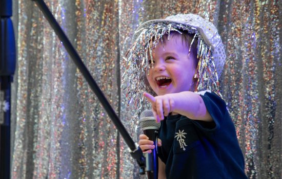 A young boy wearing a sparkly hat holds a microphone has he smiles and points. Behind him is a sequin curtain.