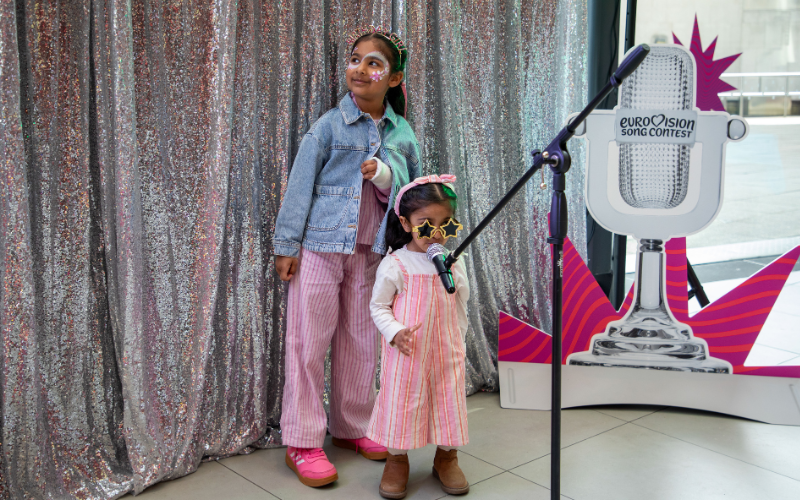 Two young girls standing in front of a sequin curtain, the youngest girl wearing pink overalls and yellow star sunglasses sings into a microphone