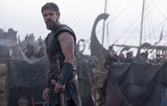 Still from The Odyssey. A man wearing traditional Ancient Greek armour stands in front of crowds of people getting boats ready to set sail.