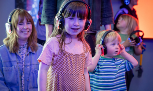 A young girl looks to camera while wearing silent disco headphones. Behind her, a young boy in a blue striped top and a woman in a demin jacket also wear headphones.