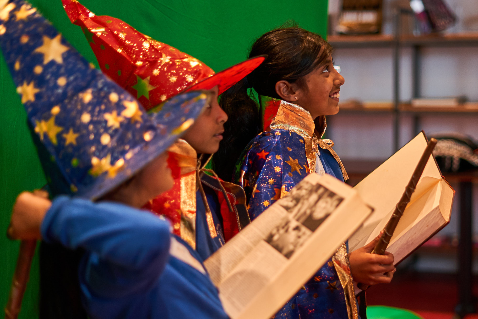 Three children dressed as wizards. Two have pointy red and blue hats with gold stars on. A girl at the front is smiling, holding a large open book and a wand.