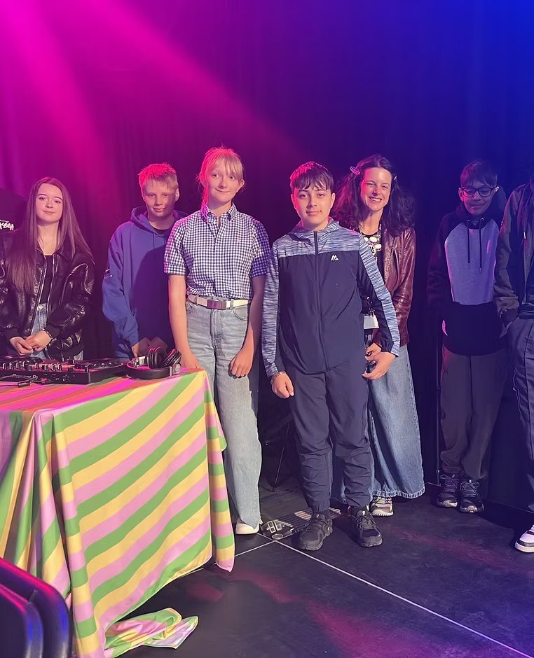 A group of young people standing on stage, smiling at the camera. On the table in front of them there is DJ equipment.