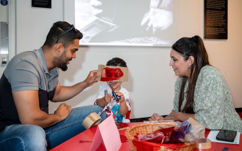 A family doing an activity which involves cutting coloured film to create filters. A dad is holding a red film over his young son's face.