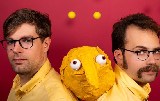 Two men in yellow outfits with a strange puppet made of tape between them