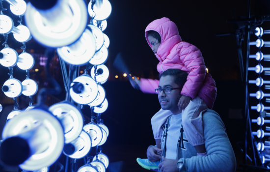 A child sitting on her dad's shoulder's hitting metal tubes surrounded by lights with a rod to make sound