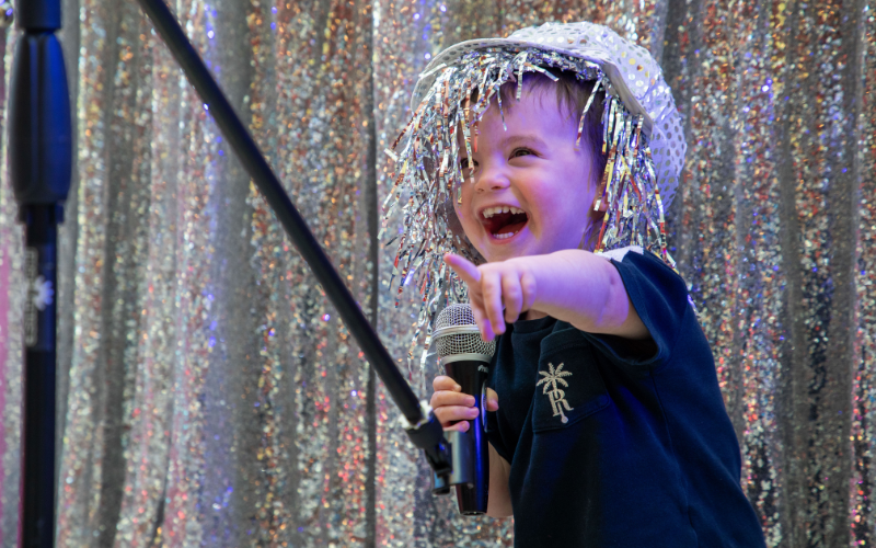 A young boy wearing a sparkly hat holds a microphone has he smiles and points. Behind him is a sequin curtain.