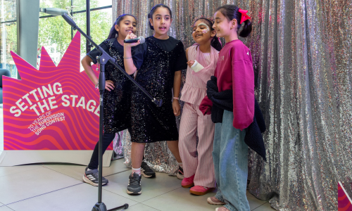 A group of four young South Asian girls pose while holding a microphone in front of a sequin curtain.
