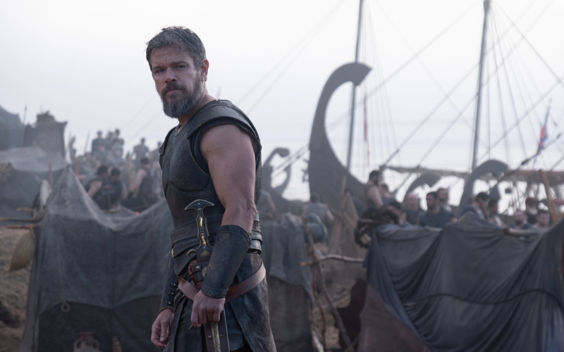Still from The Odyssey. A man wearing traditional Ancient Greek armour stands in front of crowds of people getting boats ready to set sail.