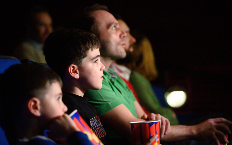Two young boys and their dad eating popcorn and looking at a cinema screen