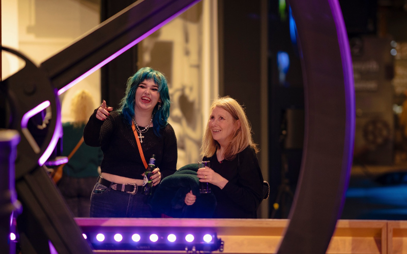 Two adults, a woman with blue hair and an older woman with blonde hair, smiling and looking at an object in a museum gallery