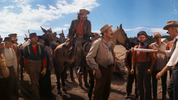 A group of men in Western attire, some on horseback, stand in a dusty landscape under a clear sky. They appear engaged in a tense discussion.