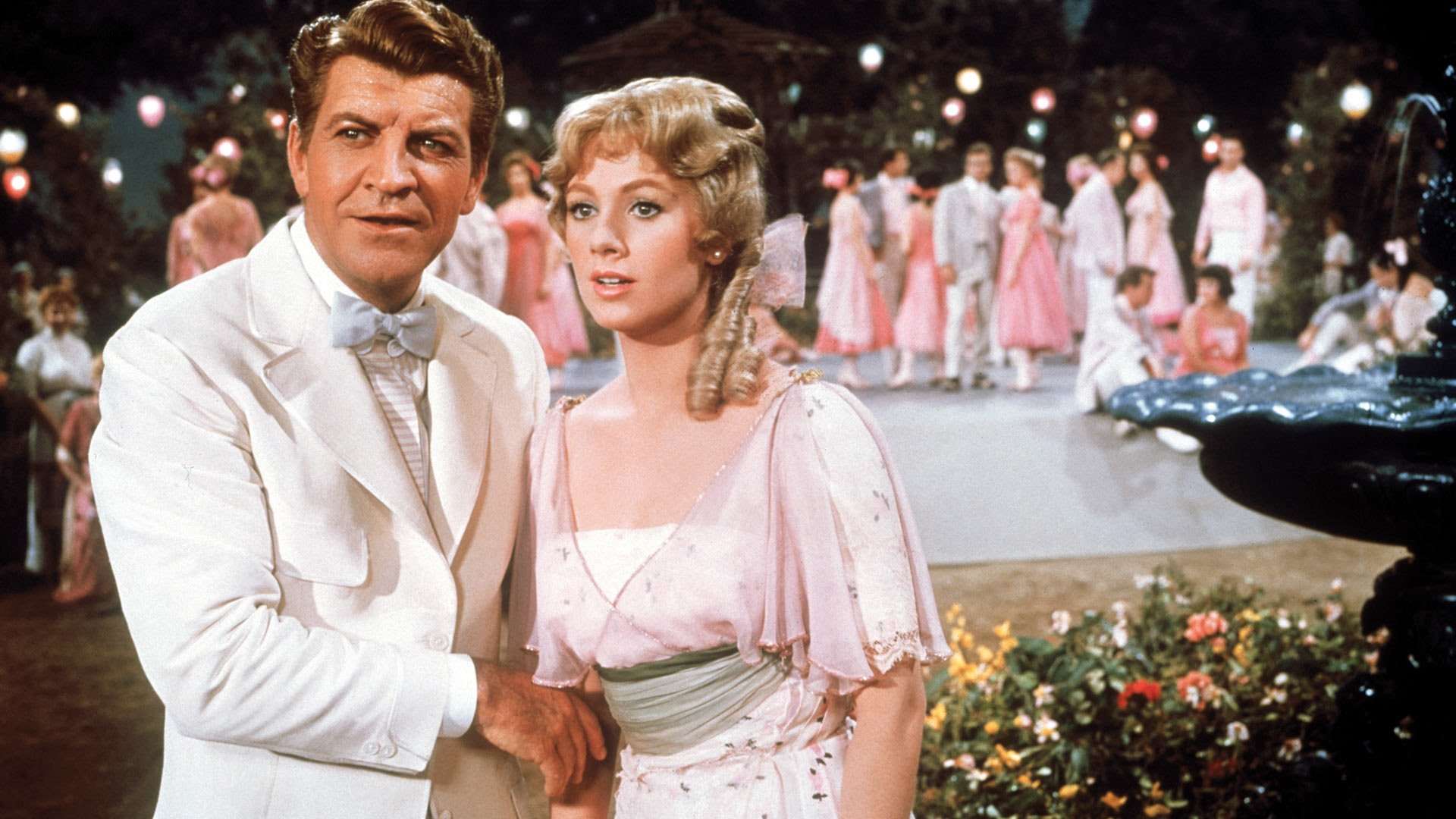 A still from The Music Man, showing a man in a white suit standing beside a young woman in a pale pink dress at an outdoor evening social, with couples dancing beneath colourful lanterns in the background.
