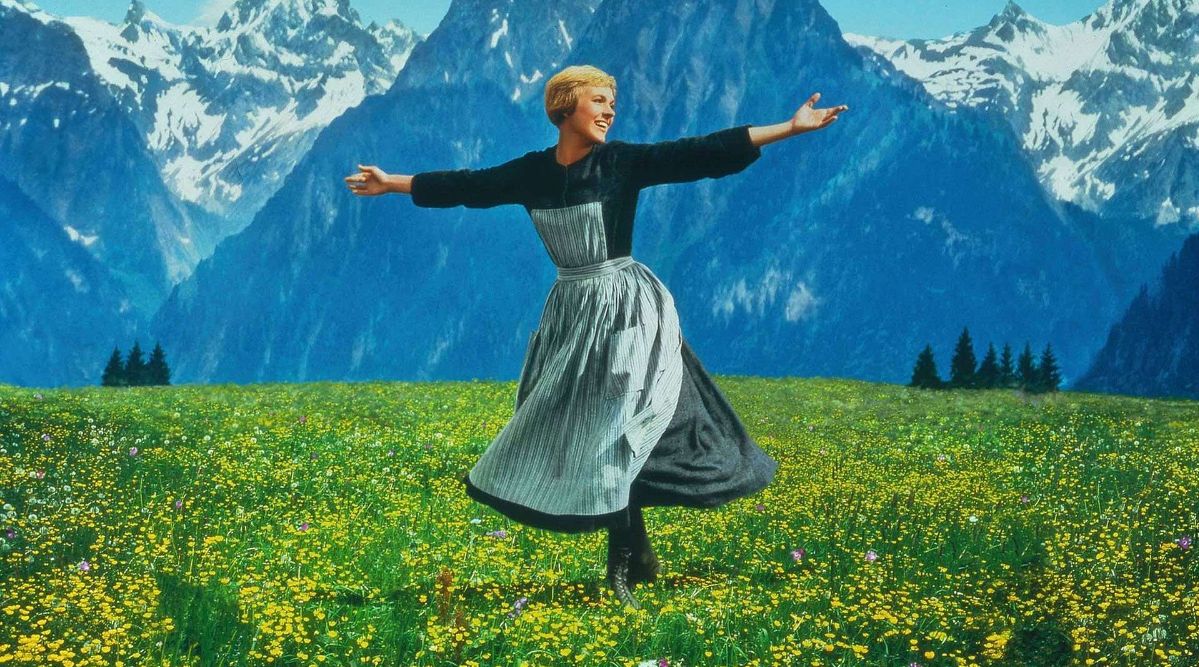 A still from The Sound of Music, showing a young woman with outstretched arms twirling in a flower-filled alpine meadow, with snow-capped mountains rising behind her.