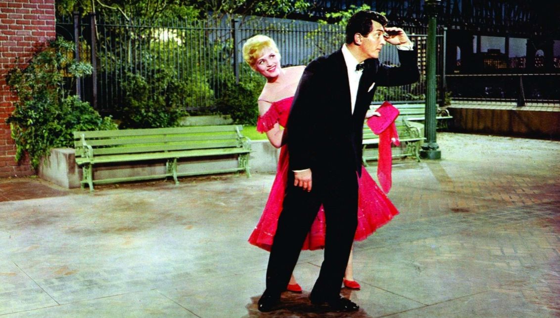 A still from Bells Are Ringing, showing a man in a black tuxedo and a woman in a bright pink off-the-shoulder dress striking a playful pose together on a nighttime city street set.