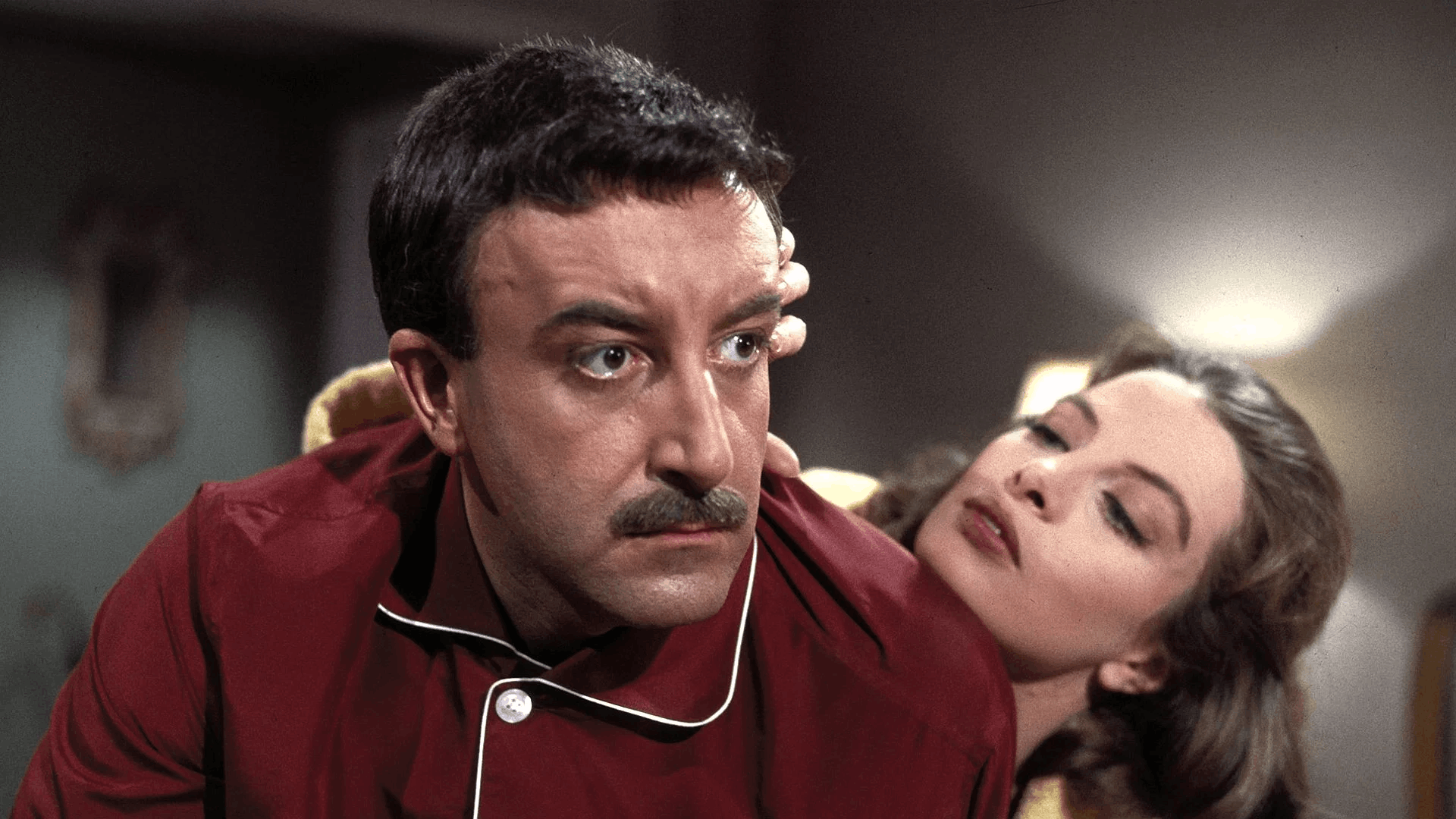 A still from The Pink Panther, a man in red pajamas looks forward with a serious expression while a woman leans closely toward him, touching his head. Indoor setting with soft lighting.