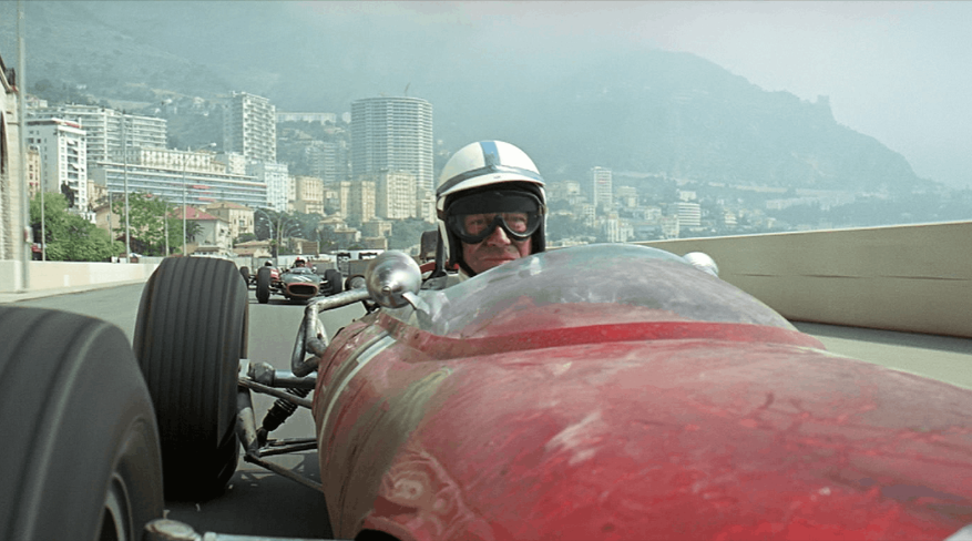 A man driving a red race car on a winding road, showcasing speed and excitement in a racing scene from Grand Prix.