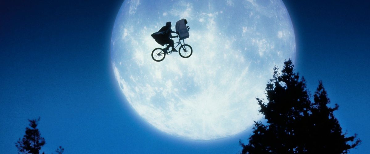 Film still from E.T. the Extra-Terrestrial showing the iconic silhouette of Elliott riding his bicycle across a massive, glowing full moon. Elliott is wearing his hooded sweatshirt, and E.T. sits wrapped in a white blanket inside the front bicycle basket. The bicycle and its riders are perfectly framed as a dark silhouette directly in front of the bright, textured white and blue moon. Dark pine tree tops line the bottom of the frame against a deep blue night sky.