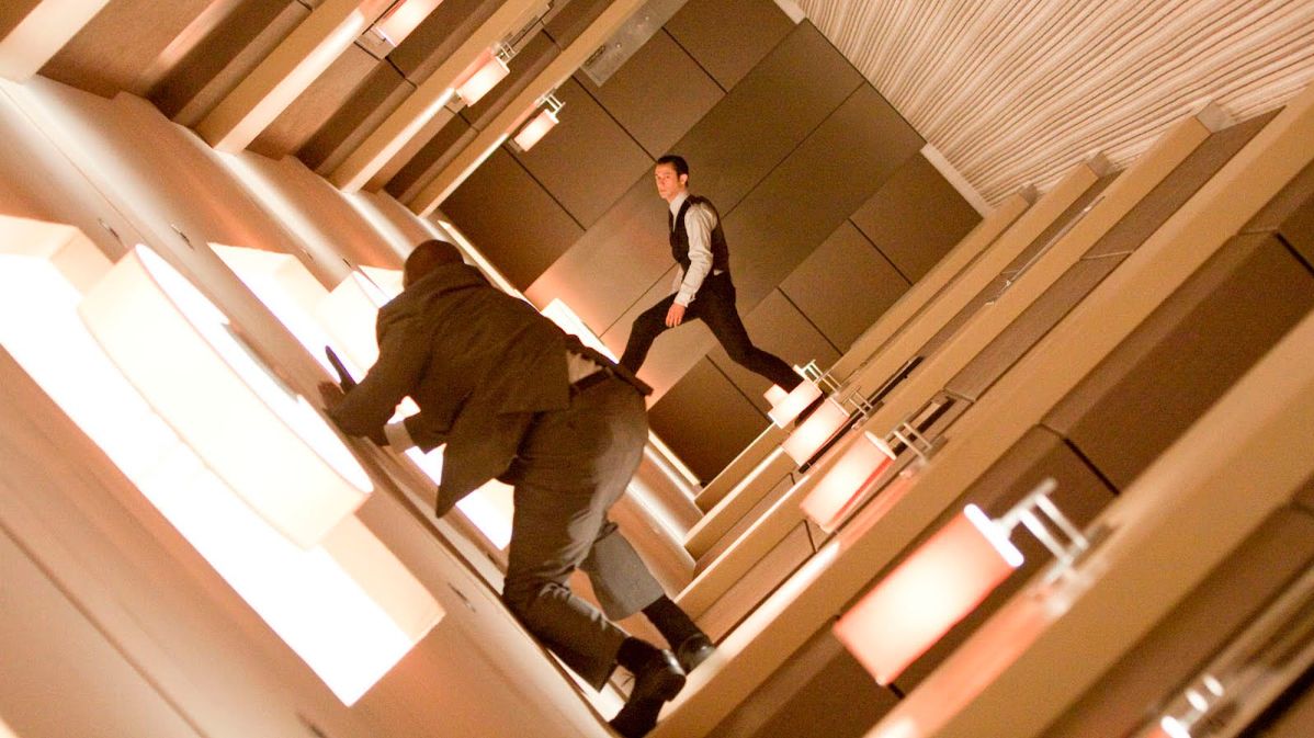 Film still from Inception showing Arthur (Joseph Gordon-Levitt) and a projection sub-security guard engaged in a zero-gravity fight inside a rotating hotel corridor. The camera is tilted significantly to the right, making the side wall with rows of glowing, rectangular light fixtures appear as the floor. Arthur is in the background, balanced mid-stride on the light panels in a white dress shirt, dark vest, and trousers. The guard is closer to the foreground, viewed from behind in a grey suit jacket and pants, leaning forward as he grips a weapon. The hallway features brown, geometric wall panels and a white, textured ceiling to the right.