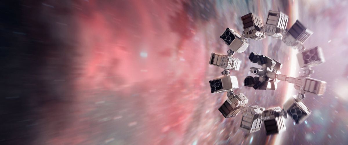 Film still from Interstellar showing the ring-shaped Endurance spacecraft spinning in deep space near a massive, colorful wormhole. The spacecraft is composed of twelve interconnected rectangular modules arranged in a perfect circle with a central docking hub. It is positioned on the right side of the frame, tilted slightly. The background features a sweeping, distorted cosmic vista filled with vibrant shades of red, orange, and blue gas clouds, creating a strong sense of motion and immense scale as light bends around the celestial anomaly.