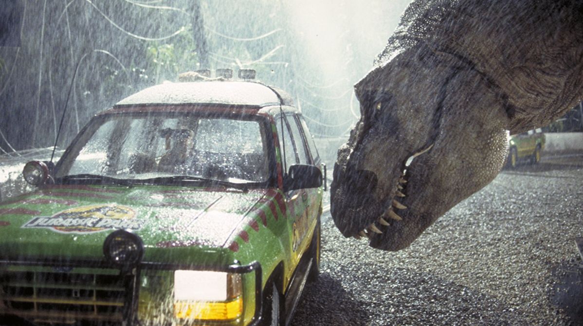 Film still from Jurassic Park showing a massive Tyrannosaurus rex leaning down next to a branded Jurassic Park Ford Explorer tour vehicle during a heavy downpour at night. The T-rex's large, detailed head is in profile on the right side of the frame, its mouth slightly open to reveal sharp teeth as it stares toward the car. The green, yellow, and red tour vehicle sits on a gravel road on the left, with rain visibly pelting the windshield and hood. The scene is intensely backlit, highlighting the thick falling rain and the textured skin of the dinosaur.