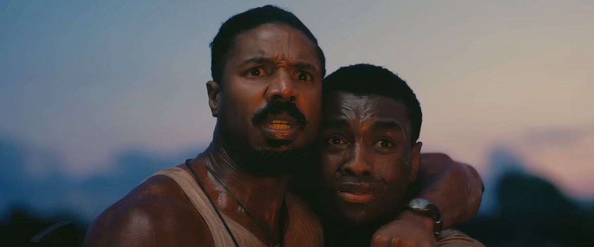 Film still from Sinners showing Smoke (Michael B. Jordan) and Sammie Moore (Miles Caton) standing close together outdoors against a dimming, twilight sky. Smoke, on the left, has a prominent mustache and goatee beard, wears a beige ribbed tank top, and looks forward with wide, terrified eyes and a tense, open-mouthed expression. Sammie, on the right, has a similarly distraught expression with a small visible scar on his forehead. Smoke has wrapped his arm tightly around Sammie's neck in a protective embrace, with his hand and watch visible near Sammie's shoulder. Both men appear sweaty and distressed under the soft evening light.