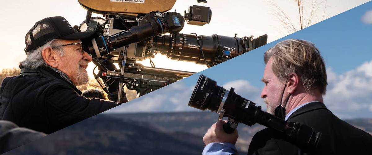 Promotional material showing a split-screen image of directors Steven Spielberg and Christopher Nolan looking through camera viewfinders on their respective movie sets. On the left half, Steven Spielberg is seen from the chest up in profile, smiling as he looks through the viewfinder of a massive Panavision film camera. He wears a black baseball cap, glasses, and a black jacket under warm, golden hour sunlight. On the right half, Christopher Nolan is shown from the chest up in profile against a blue sky with soft white clouds, holding a handheld director's viewfinder to his eye. He wears a dark suit jacket and has a black face mask pulled down under his chin. A diagonal dark line splits the two scenes down the center.
