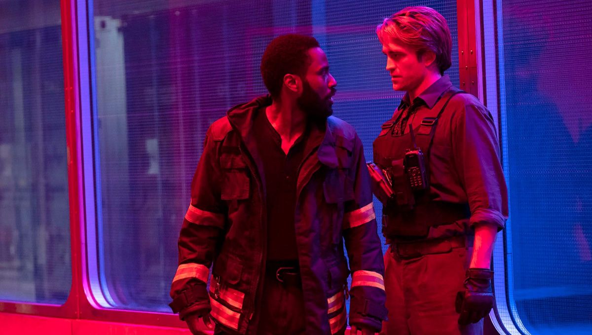 Film still from Tenet showing the Protagonist (John David Washington) and Neil (Robert Pattinson) standing close together and looking intensely at each other inside a room split by a large glass window. The room is heavily saturated with deep red lighting on their side, while the area visible through the glass windows behind them is illuminated by a stark blue light. The Protagonist wears a dark jacket with reflective orange strips, and Neil wears a dark tactical vest with a walkie-talkie attached to his chest.