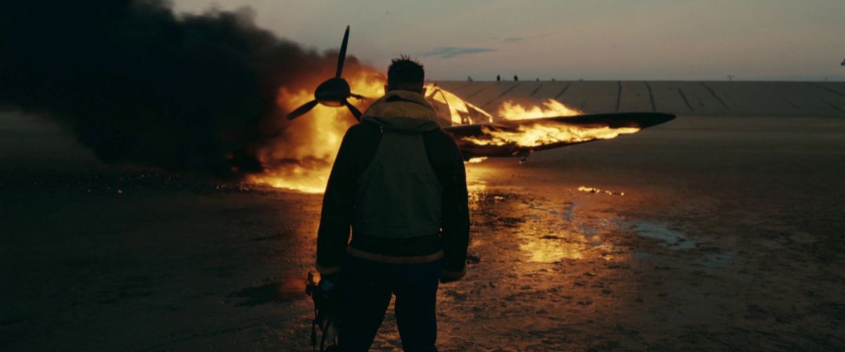 Film still from Dunkirk showing Royal Air Force pilot Farrier (Tom Hardy) from behind, standing on a vast beach at dusk as his fighter plane burns intensely in front of him. Farrier wears a dark flight jacket with a cream-colored shearling collar and a tactical vest. Thick black smoke billows from the aircraft as bright orange flames consume the cockpit and wings, casting a strong golden reflection across the damp sand. In the distant background, a low concrete seawall or ridge stretches across the horizon under a dim evening sky.