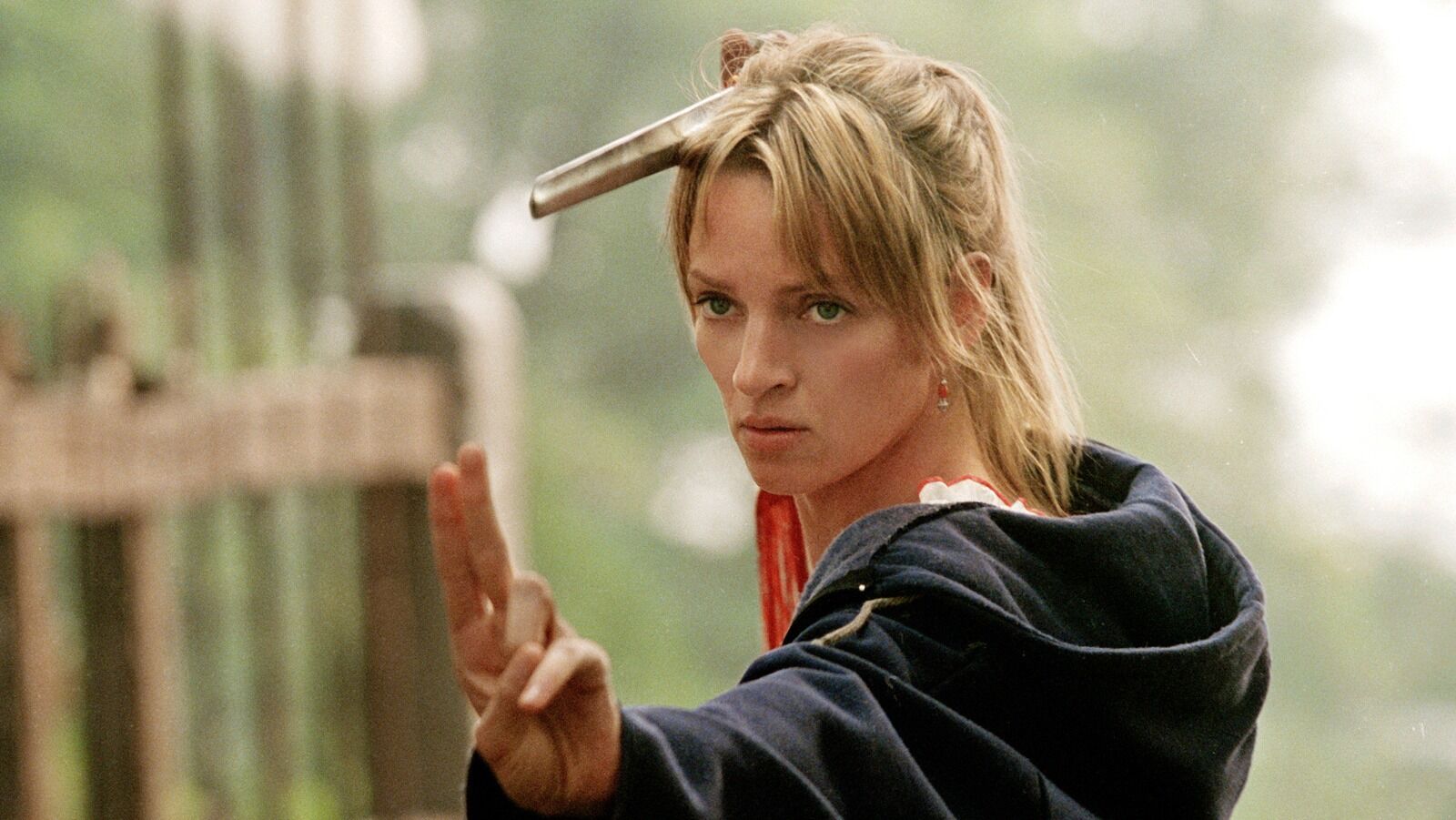 A woman with a focused expression holds two fingers up in a martial arts stance, wearing a blue robe, with a sword slung over their shoulder, outdoors.