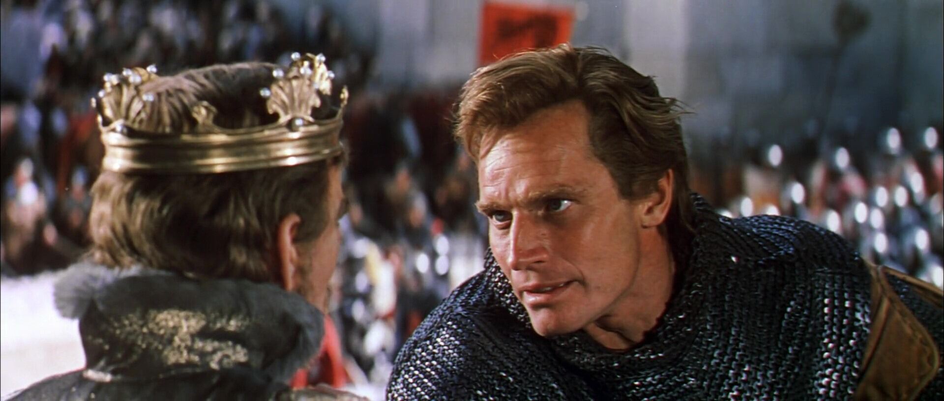 A man in chainmail armor intensely confronts a crowned figure. The background shows a blurry crowd, conveying a medieval setting and high tension.