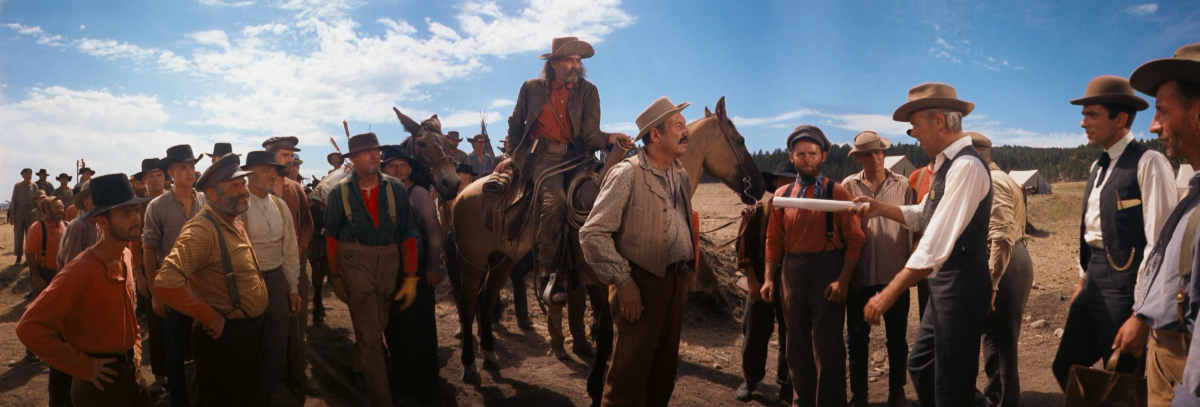 A group of men in Western attire, some on horseback, stand in a dusty landscape under a clear sky. They appear engaged in a tense discussion.
