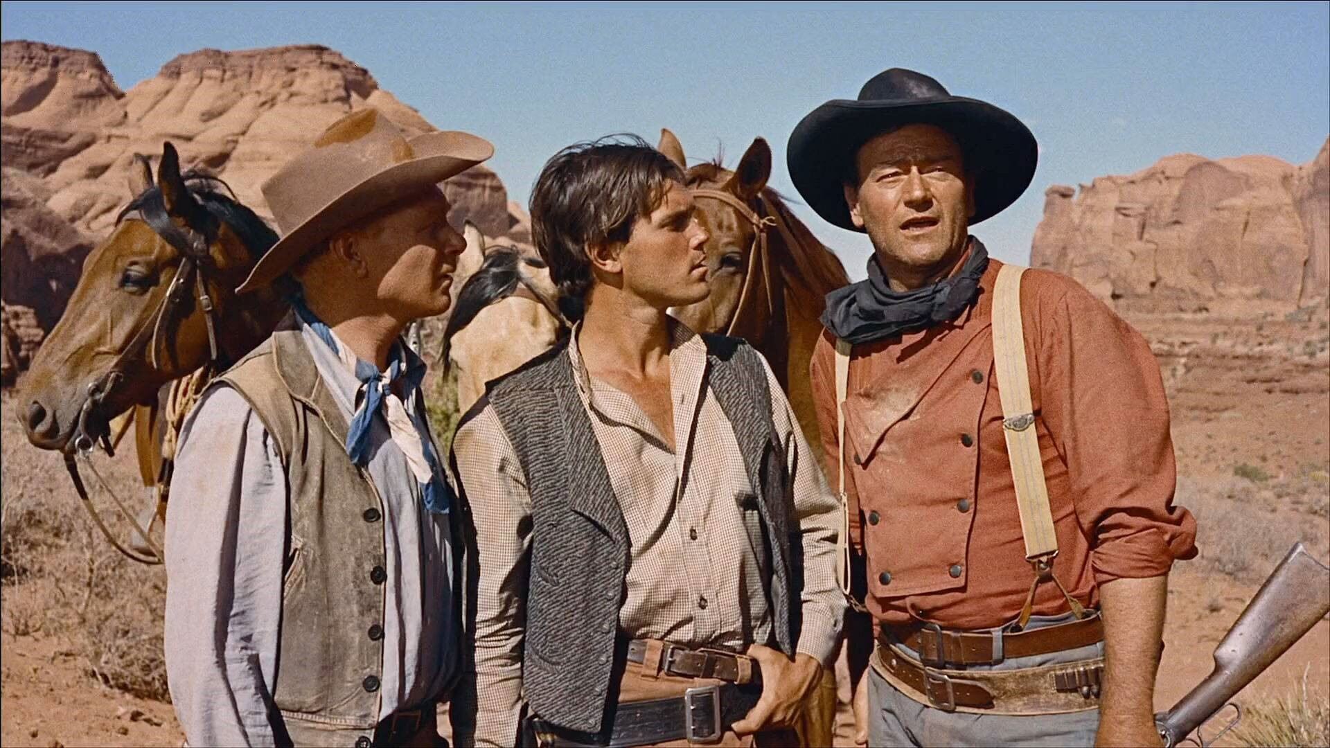 Three cowboys stand in a rocky desert landscape with two horses. The men wear hats and vests, looking intently ahead, conveying a serious tone.