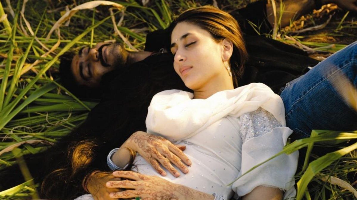A couple lies in lush green grass. The woman in white, eyes closed, looks serene, with henna on her hands. The man beside her appears relaxed.
