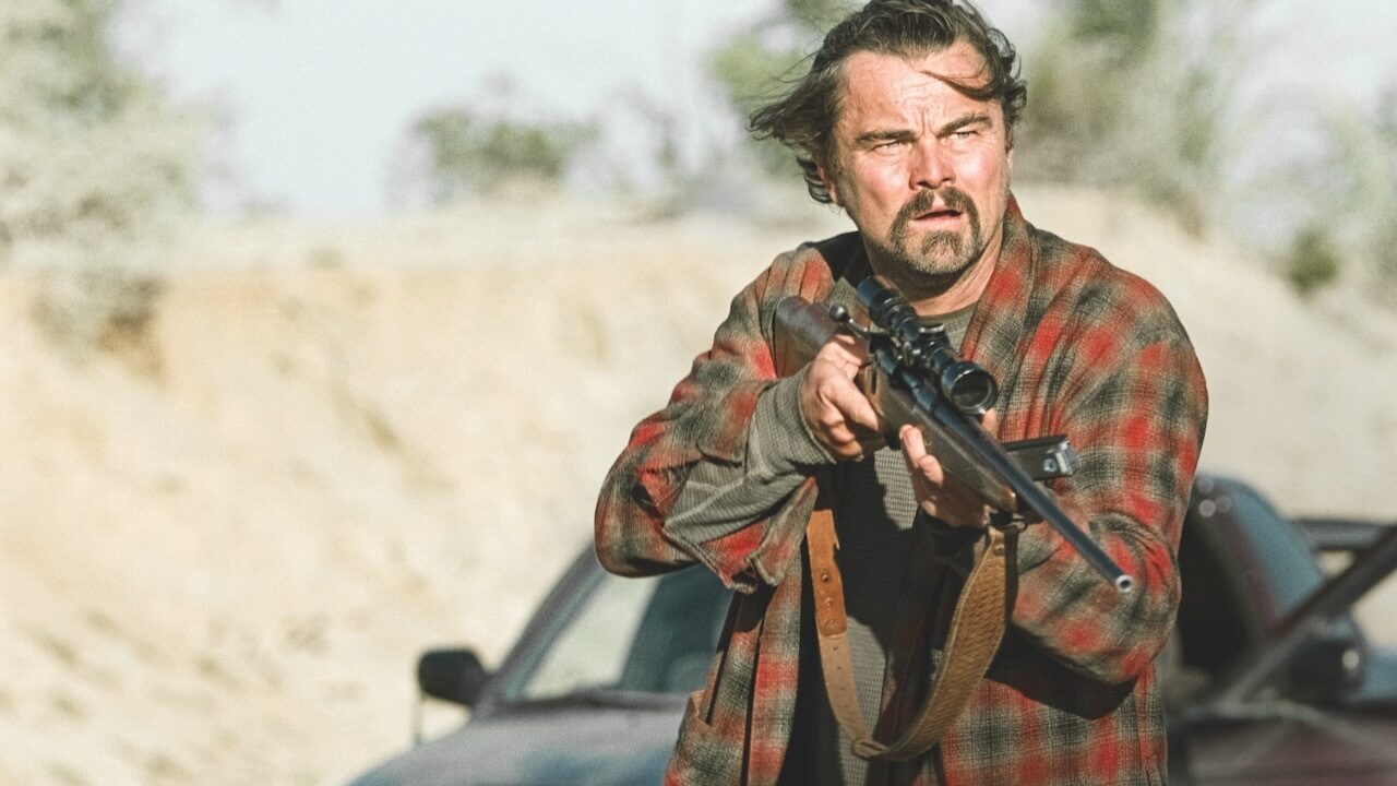 Man in rugged landscape, holding a rifle, looks focused and intense. Wearing a red plaid shirt, he stands near a black vehicle with a blurred background.