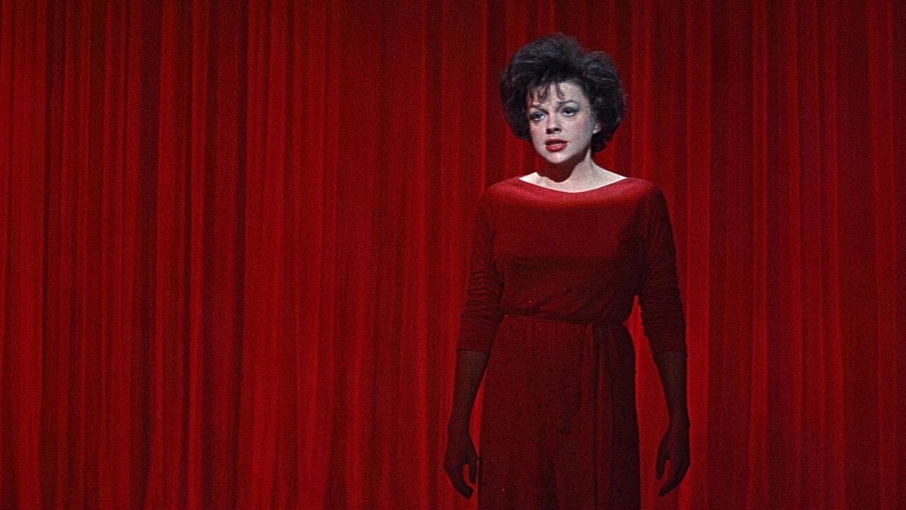 A woman in a red dress stands against lush red curtains, exuding a dramatic and intense mood. Her expression is serious, adding to the theatrical atmosphere.