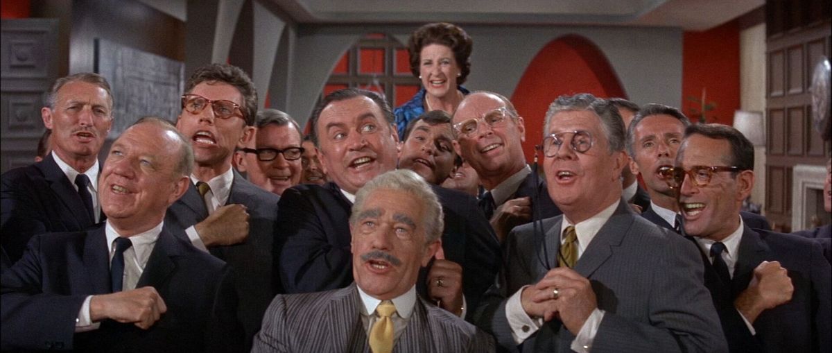 A group of men in suits enthusiastically sing together, their expressions joyous and animated. A smiling woman stands behind them, adding to the celebratory mood.