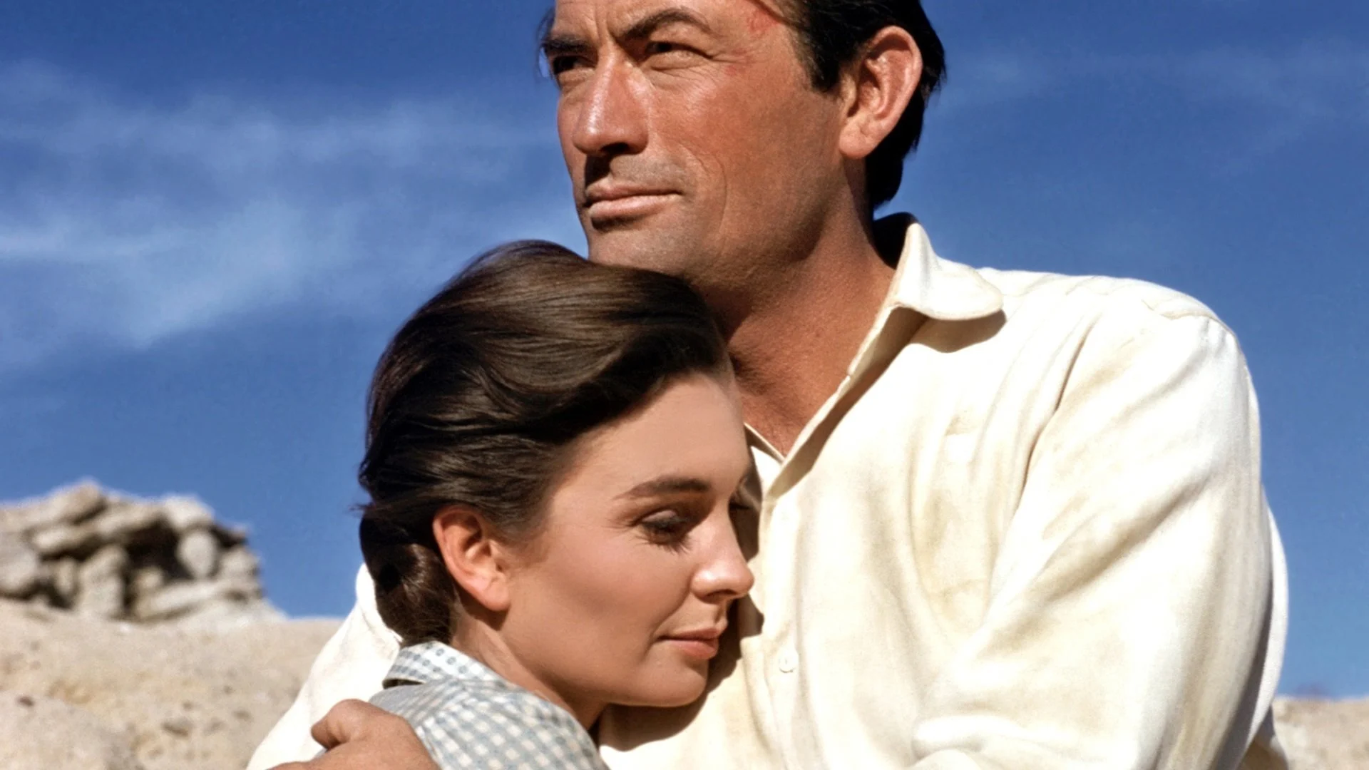 A couple embraces against a clear blue sky. The man looks thoughtful, while the woman rests her head on his chest, eyes closed, conveying warmth and serenity.