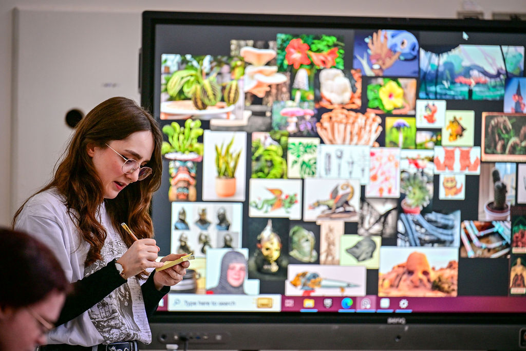A woman with hark dair and glasses stands in front of a screen showing exmaple sof artistic prompts.