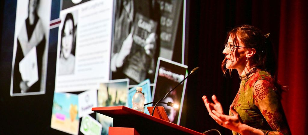 A woman with glasses and pulled back hair speaks at a podium with a presentation on a large screen behind her.