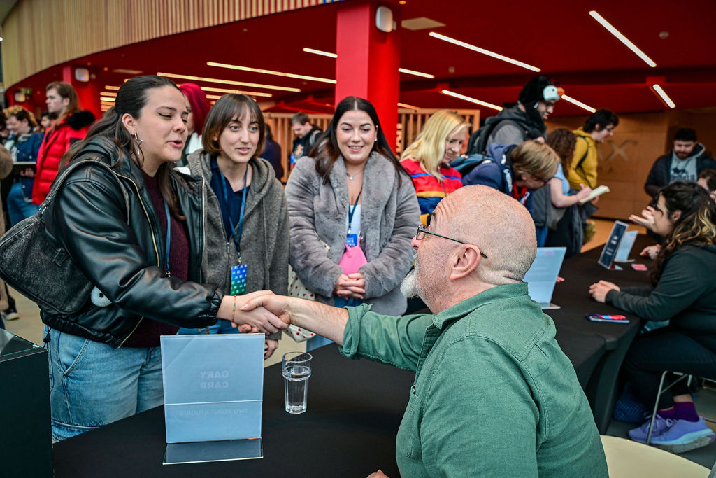 A man sat wearing a green shirt shsakes hands with a young woman stood wearing a leather jacket while two other young women stand adjacently.
