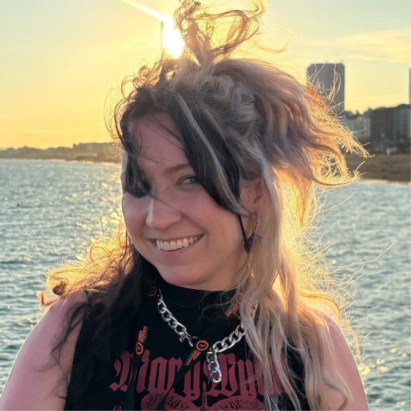 Person smiling by the water at sunset, with windblown two-tone hair, wearing a black graphic top and chain necklace, city buildings visible in the background.