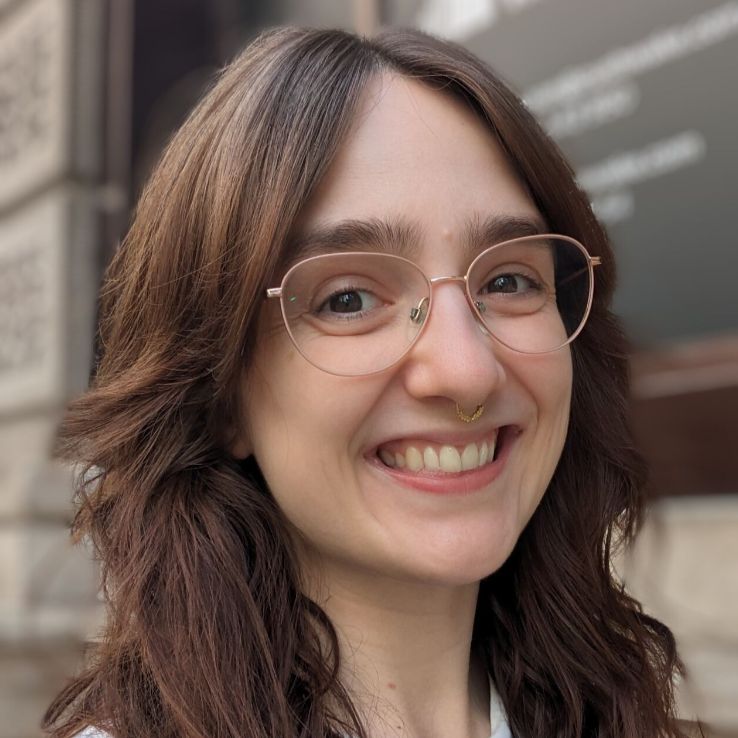 Smiling person with long brown wavy hair wearing round glasses and a small septum ring, standing outdoors in front of a stone building.