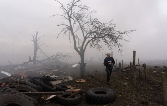 A journalist walks through a ruined landscape in which only bare trees remain standing amidst the rubble.